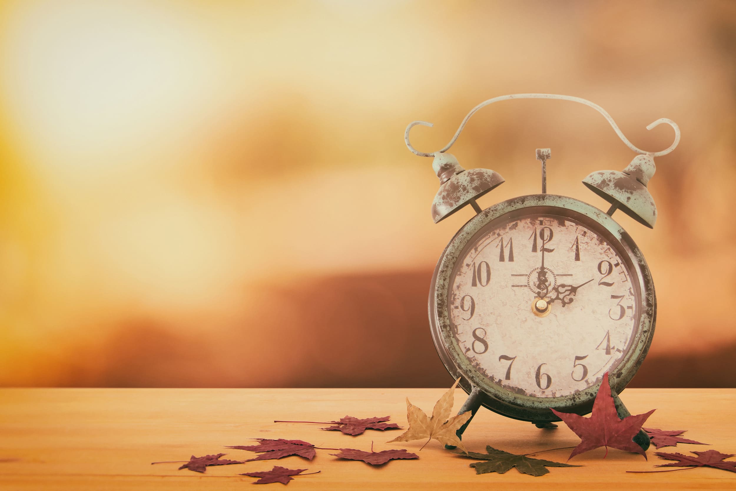 antique alarm clock on table surrounding by fall leaves with sunlight shining in the background