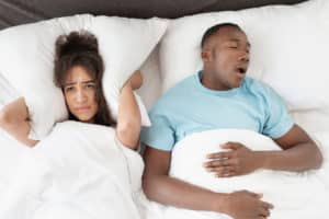couple in bed, man is snoring, woman covers her ears with pillow
